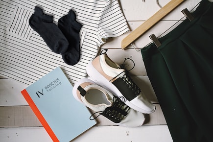 A flat lay arrangement featuring a striped shirt, a pair of black socks, a skirt on a hanger, a pair of stylish sneakers, and a book titled 'Invictus Tailoring' placed on a white wooden surface.