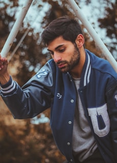 A man is wearing a letterman jacket and holding onto a pole. He looks downward with a contemplative expression. The background features blurred brown foliage, suggesting an outdoor setting.