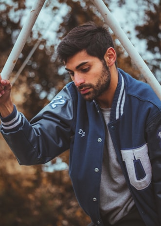 A man is wearing a letterman jacket and holding onto a pole. He looks downward with a contemplative expression. The background features blurred brown foliage, suggesting an outdoor setting.