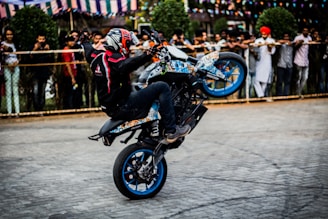 A motorcyclist performs a wheelie on a colorful bike in front of a crowd. The rider wears a helmet and protective gear, and the onlookers are behind a fence, watching the stunt. Decorative flags are strung in the background.
