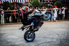 A motorcyclist performs a wheelie on a colorful bike in front of a crowd. The rider wears a helmet and protective gear, and the onlookers are behind a fence, watching the stunt. Decorative flags are strung in the background.