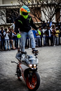A person wearing a bright multicolored helmet and jacket performs a stunt by standing on a moving motorcycle. The bike has orange and white bodywork, and the person is balancing on one leg with their arms extended. Spectators in casual attire watch from behind a barrier.