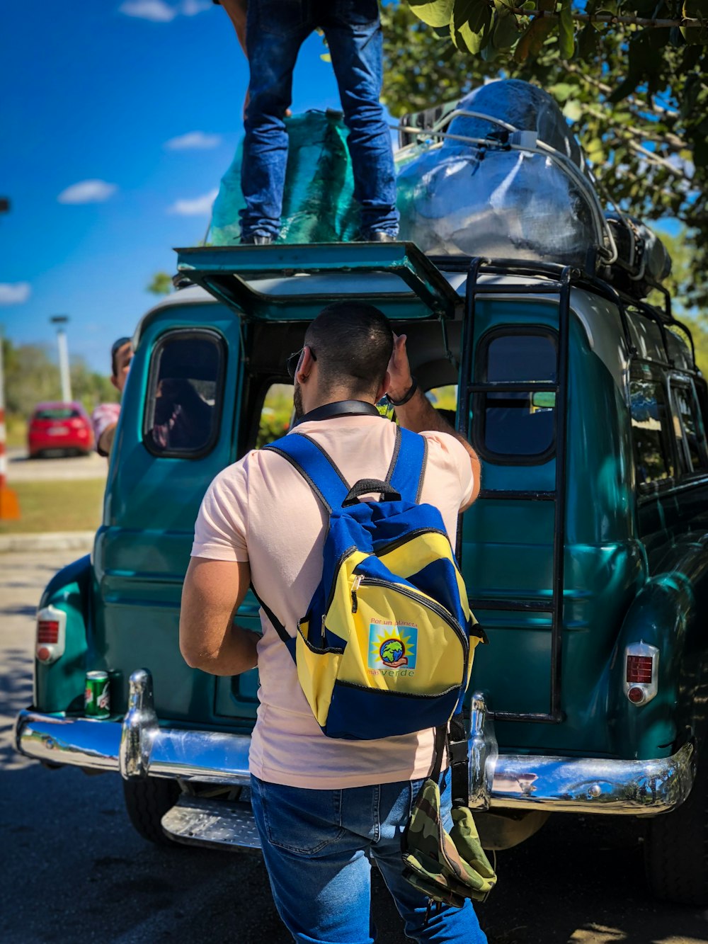 man in white shirt with yellow and blue backpack standing at back of vehicle