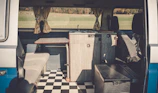 The interior of a camper van features a compact kitchen setup, including a small wooden counter and a built-in sink. Opposite the kitchen, there's a cushioned bench seat. The floor is covered with a black and white checkered pattern, and light is streaming in through curtained windows, providing a view of a grassy landscape outside.