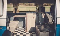The interior of a camper van features a compact kitchen setup, including a small wooden counter and a built-in sink. Opposite the kitchen, there's a cushioned bench seat. The floor is covered with a black and white checkered pattern, and light is streaming in through curtained windows, providing a view of a grassy landscape outside.