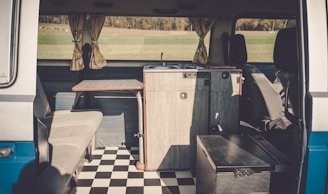 The interior of a camper van features a compact kitchen setup, including a small wooden counter and a built-in sink. Opposite the kitchen, there's a cushioned bench seat. The floor is covered with a black and white checkered pattern, and light is streaming in through curtained windows, providing a view of a grassy landscape outside.