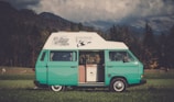 A classic teal and white camper van is parked on a lush green field with mountains in the background. The van features graphics and text on its roof, and its side door is open to reveal cabinets inside. Cows can be seen grazing in the field, adding to the serene rural setting. The sky is partly cloudy, casting a soft light over the scene.