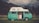 A classic teal and white camper van is parked on a lush green field with mountains in the background. The van features graphics and text on its roof, and its side door is open to reveal cabinets inside. Cows can be seen grazing in the field, adding to the serene rural setting. The sky is partly cloudy, casting a soft light over the scene.