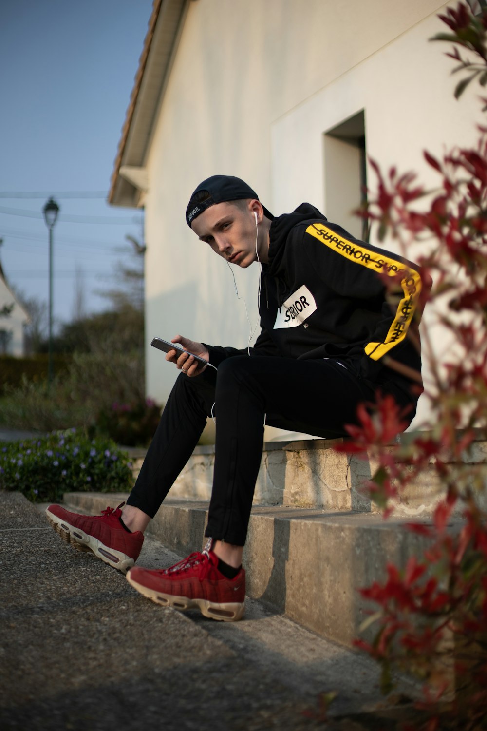 Hombre con sudadera con capucha negra y pantalones sentado junto a las escaleras escuchando en su teléfono inteligente