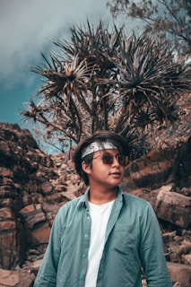 A person wearing sunglasses, a headband, and a teal button-up shirt over a white t-shirt is standing outdoors. The background features rocky terrain and a spiky tree or bush, with a partly cloudy sky overhead.