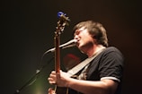 A musician passionately sings into a microphone while playing an acoustic guitar. The stage lighting creates a warm, intimate atmosphere with a slight focus on the musician.
