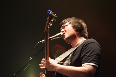 A musician passionately sings into a microphone while playing an acoustic guitar. The stage lighting creates a warm, intimate atmosphere with a slight focus on the musician.