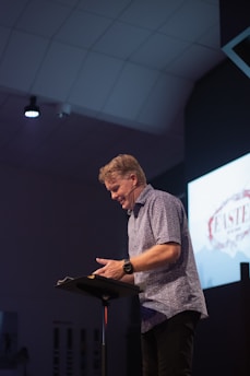 A person is standing at a podium in a dimly lit indoor setting, reading or speaking. They are wearing a patterned short-sleeve shirt and are positioned slightly to the side, facing the podium. A screen in the background displays a graphic that includes the word 'Easter'. The ceiling has a grid-like pattern and a spotlight is visible.