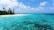 A picturesque tropical beach scene with crystal clear turquoise water, white sandy shore, and lush palm trees along the coastline. The horizon shows a blue sky with scattered white clouds and a series of small overwater bungalows in the distance.