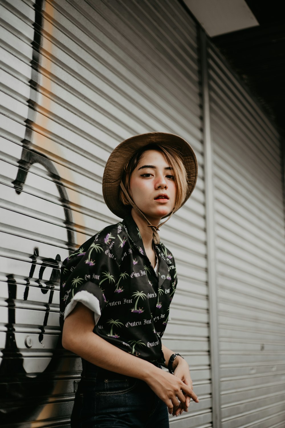 woman in black button-up shirt and brown hat elaning on roll-up door