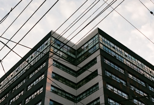 A multi-story building with numerous windows, viewed from a low angle. There are multiple electrical wires crossing the frame diagonally.