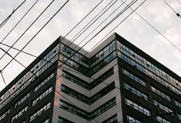 A multi-story building with numerous windows, viewed from a low angle. There are multiple electrical wires crossing the frame diagonally.
