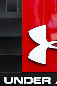 A large, bold logo featuring the brand 'Under Armour' is prominently displayed. The design includes a distinctive white emblem resembling an abstract 'U' and 'A' intertwined, set against a backdrop of black and red panels. The word 'UNDER' is written in capital letters below the logo in white.