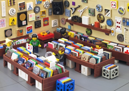 A scene depicting a LEGO brick-built record store with colorful records and artwork on the walls. Three LEGO minifigures are interacting within the store, browsing and discussing the records. The walls are covered with various colorful tiles and artistic designs, giving the store a vibrant and eclectic atmosphere. The store has wooden shelves and displays a vintage vibe with its diverse decor.