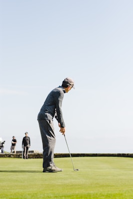 A golfer in a cap and casual sporting attire is preparing to putt on the green of a golf course. The golf course stretches out under a clear blue sky, with other players and a golf cart visible in the background.