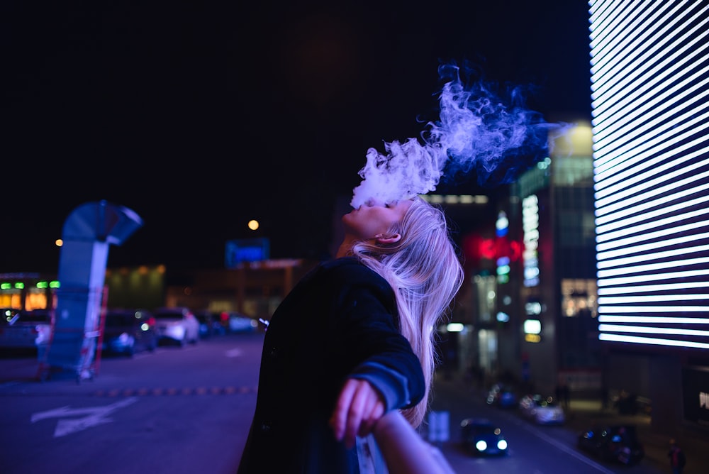 closed eyed woman leaning backwards on handrail blowing smoke at the city during night