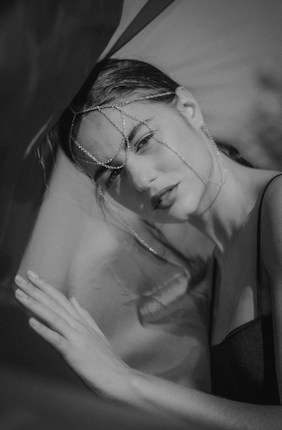 A monochrome portrait of a woman wearing a delicate chain headpiece. Her expression is pensive, and she gazes directly at the camera. She rests her hand gently against a surface, partially obscured by sheer fabric, creating an ethereal effect.