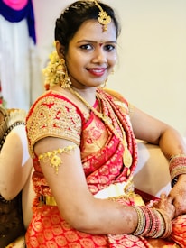 A woman dressed in traditional attire featuring a richly embroidered red saree with gold jewelry and floral adornments. She is seated on a cushioned seat, smiling warmly with henna designs on her hands. The background includes soft, colorful fabric decorations.