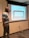 A man stands in front of a projection screen in a classroom or meeting room. He points to the screen, which displays an image of a table with the text 'In groups or whole group discuss: What's on the table?'. The room has beige walls, a whiteboard, and visible electrical outlets and cords on the floor.