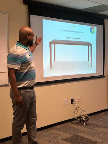 A man stands in front of a projection screen in a classroom or meeting room. He points to the screen, which displays an image of a table with the text 'In groups or whole group discuss: What's on the table?'. The room has beige walls, a whiteboard, and visible electrical outlets and cords on the floor.