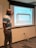 A man stands in front of a projection screen in a classroom or meeting room. He points to the screen, which displays an image of a table with the text 'In groups or whole group discuss: What's on the table?'. The room has beige walls, a whiteboard, and visible electrical outlets and cords on the floor.
