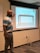A man stands in front of a projection screen in a classroom or meeting room. He points to the screen, which displays an image of a table with the text 'In groups or whole group discuss: What's on the table?'. The room has beige walls, a whiteboard, and visible electrical outlets and cords on the floor.
