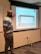 A man stands in front of a projection screen in a classroom or meeting room. He points to the screen, which displays an image of a table with the text 'In groups or whole group discuss: What's on the table?'. The room has beige walls, a whiteboard, and visible electrical outlets and cords on the floor.