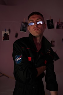 A person wearing a NASA-themed black jumpsuit and reflective sunglasses stands in a dimly lit room. The background features a string with photos clipped onto it. The lighting casts dark shadows, and there is a hint of red light illuminating the scene.