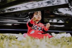 A young child is sitting on the grass, wearing a red racing-themed outfit with black and white checkered patterns. The child is holding a small object and is positioned in front of a black vehicle.