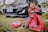 A child is joyfully sitting on a red push car, dressed in a red racing suit with checkerboard patterns. The scene is outdoors on grass with a black car parked behind and toys scattered around.