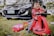A child is joyfully sitting on a red push car, dressed in a red racing suit with checkerboard patterns. The scene is outdoors on grass with a black car parked behind and toys scattered around.