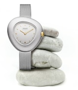 A sleek wristwatch with a metallic mesh band and a uniquely shaped, asymmetrical face is elegantly displayed. The watch features a silver casing, a simple dial with gold accents, and a single gold crown. It is artfully balanced on a stack of smooth, round gray stones, creating a minimalist and modern aesthetic.
