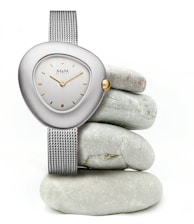 A sleek wristwatch with a metallic mesh band and a uniquely shaped, asymmetrical face is elegantly displayed. The watch features a silver casing, a simple dial with gold accents, and a single gold crown. It is artfully balanced on a stack of smooth, round gray stones, creating a minimalist and modern aesthetic.