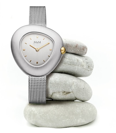 A sleek wristwatch with a metallic mesh band and a uniquely shaped, asymmetrical face is elegantly displayed. The watch features a silver casing, a simple dial with gold accents, and a single gold crown. It is artfully balanced on a stack of smooth, round gray stones, creating a minimalist and modern aesthetic.