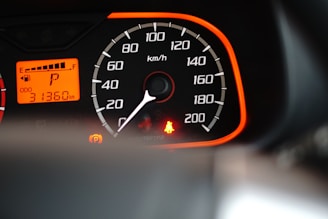 A close-up view of a car speedometer and odometer with an orange backlit panel. The speedometer scale is marked from 0 to 200 km/h, and the needle points to zero. The odometer reads 31,360 kilometers. There is a bright red seatbelt warning light and a parking indicator light illuminated.