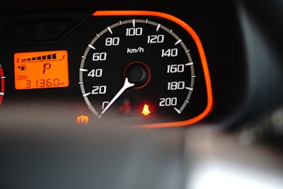 A close-up view of a car speedometer and odometer with an orange backlit panel. The speedometer scale is marked from 0 to 200 km/h, and the needle points to zero. The odometer reads 31,360 kilometers. There is a bright red seatbelt warning light and a parking indicator light illuminated.