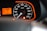 A close-up view of a car speedometer and odometer with an orange backlit panel. The speedometer scale is marked from 0 to 200 km/h, and the needle points to zero. The odometer reads 31,360 kilometers. There is a bright red seatbelt warning light and a parking indicator light illuminated.