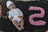 A baby lies on a dark fabric backdrop, wearing a colorful outfit with a white hat embellished with small designs. To the right of the baby, the number five is crafted from pink and white fluffy material, adding a playful element to the scene. The baby appears to be smiling, conveying a sense of joy.