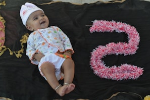 A baby lies on a dark fabric backdrop, wearing a colorful outfit with a white hat embellished with small designs. To the right of the baby, the number five is crafted from pink and white fluffy material, adding a playful element to the scene. The baby appears to be smiling, conveying a sense of joy.