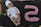 A baby lies on a dark fabric backdrop, wearing a colorful outfit with a white hat embellished with small designs. To the right of the baby, the number five is crafted from pink and white fluffy material, adding a playful element to the scene. The baby appears to be smiling, conveying a sense of joy.