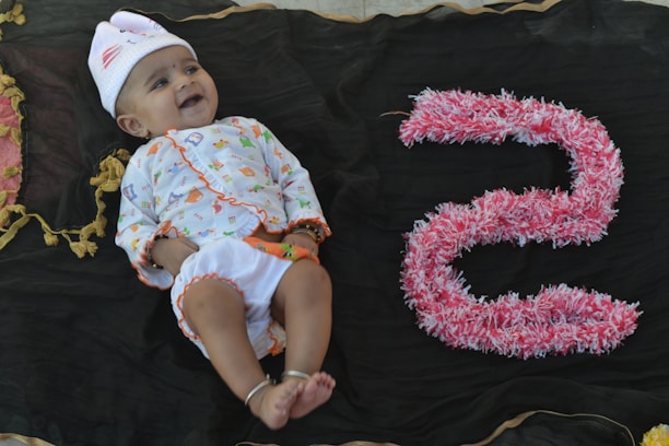 A baby lies on a dark fabric backdrop, wearing a colorful outfit with a white hat embellished with small designs. To the right of the baby, the number five is crafted from pink and white fluffy material, adding a playful element to the scene. The baby appears to be smiling, conveying a sense of joy.