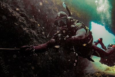 A scuba diver is exploring an underwater cave, wearing full diving gear including a mask and oxygen tank. The cave is dark with some light filtering through an opening, casting a blue hue. Another diver can be seen in the background.