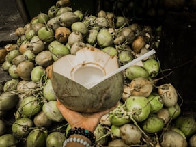 A hand is holding a partially cut, fresh coconut with a straw inserted, surrounded by a large pile of coconuts in various shades of green and brown. The hand is adorned with colorful beaded bracelets.
