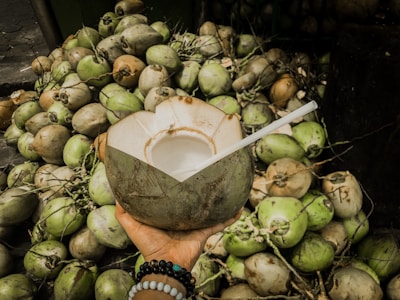 A hand is holding a partially cut, fresh coconut with a straw inserted, surrounded by a large pile of coconuts in various shades of green and brown. The hand is adorned with colorful beaded bracelets.