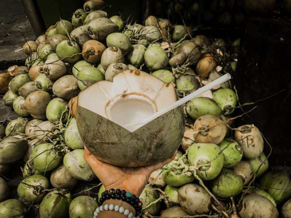 A hand is holding a partially cut, fresh coconut with a straw inserted, surrounded by a large pile of coconuts in various shades of green and brown. The hand is adorned with colorful beaded bracelets.